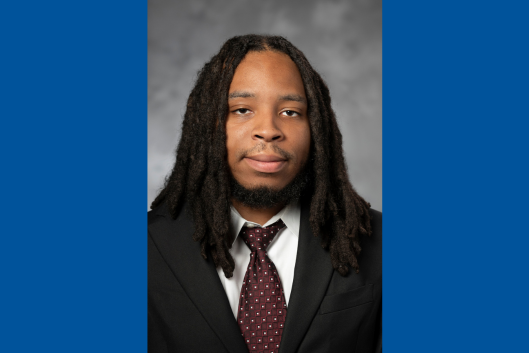 A photo of Christian Richardson, he is smiling at the camera, he has dark hair, wearing a dark suit jacket, white shirt and burgundy tie